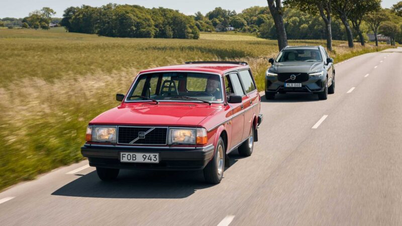 A red vintage Volvo station wagon drives ahead of a modern gray Volvo SUV on a two-lane rural road lined with trees and fields, hinting at Volvo’s evolution as it expands U.S. operations with XC60 production from 2026.