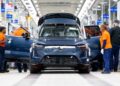 Factory workers assemble a dark blue SUV on a brightly lit automotive production line, as Volvo expands U.S. operations with XC60 production from 2026; the vehicle’s doors and hood are open during assembly.