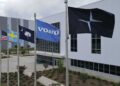Five flags—American, Swedish, South Carolina, Volvo, and Polestar—fly outside a modern industrial building on a cloudy day as Volvo expands U.S. operations with XC60 production from 2026.