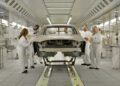 A group of people in white coveralls inspect a car frame in a brightly lit automotive manufacturing facility, as Volvo expands U.S. operations with XC60 production from 2026.