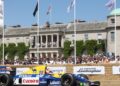 A blue and yellow Formula 1 race car, one of the 10 Fastest Cars At The 2025 Goodwood Festival Of Speed, drives past a crowd and sponsor banners in front of a large historic mansion.