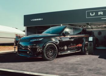 A black Urban Automotive XRS Widetrack SUV with sporty detailing and decals is displayed on a platform at an outdoor auto event at Goodwood, with branded signage in the background.