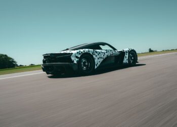 A Zenvo sports car with a black and white camouflage wrap drives at speed on a clear, open road under a blue sky, likely testing ahead of Monterey Car Week as part of the Aurora Development program.