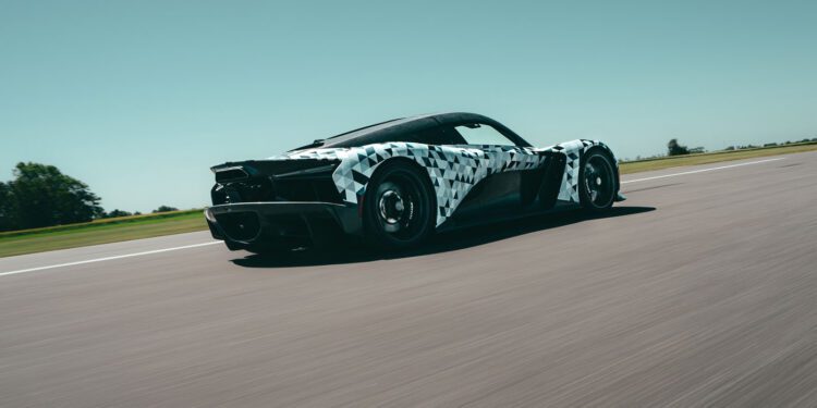 A Zenvo sports car with a black and white camouflage wrap drives at speed on a clear, open road under a blue sky, likely testing ahead of Monterey Car Week as part of the Aurora Development program.