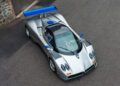 A silver Pagani sports car with a blue rear wing is parked on an asphalt surface next to a stone and brick building, ready to impress at the 2025 Monterey Car Week.