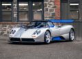 A silver Pagani sports car with blue accents and a large rear wing is parked on pavement in front of a stone and brick building, ready to impress at the 2025 Monterey Car Week.