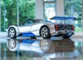 A silver and blue Pagani sports car with a large rear wing is parked indoors on a reflective floor, with trees visible through large windows in the background—an impressive sight fit for 2025 Monterey Car Week.