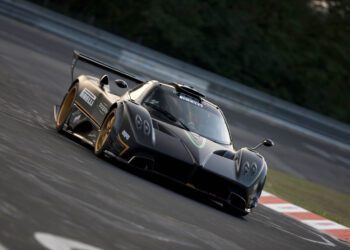 A black Pagani sports car with gold accents speeds through a racetrack corner at 2025 Monterey Car Week, slightly tilted as blurred trees and barriers rush by in the background.