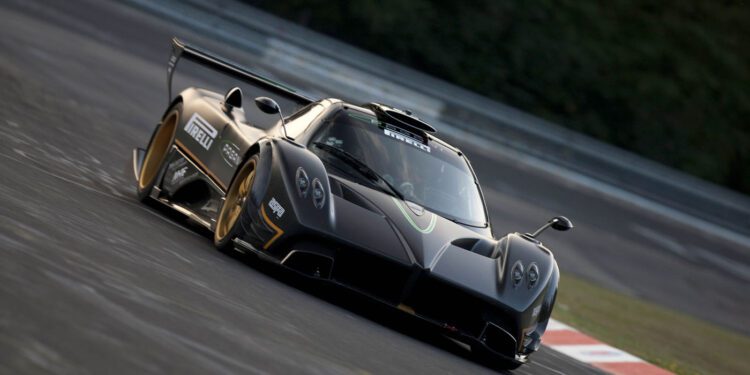 A black Pagani sports car with gold accents speeds through a racetrack corner at 2025 Monterey Car Week, slightly tilted as blurred trees and barriers rush by in the background.