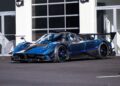 A blue and black Pagani sports car with a large rear wing is parked in front of a modern building with glass doors and windows, ready to impress at the 2025 Monterey Car Week.