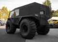A large, matte-black Atlas ATV with oversized tires, built for rugged terrain and amphibious adventures, is parked outside among other cars on a sunny day—now For Sale.