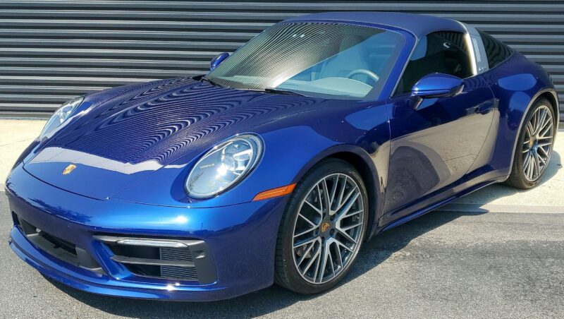 A blue Porsche 911 sports car, available with some of the best lease deals, is parked on a concrete surface in front of a black metal garage door.
