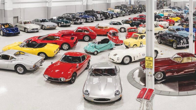 A large indoor showroom displays a diverse collection of classic and vintage cars in various colors and models, arranged in neat rows on a polished floor.
