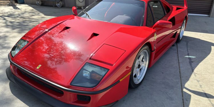 A red Ferrari F40 sports car is parked on a concrete driveway near a building with a closed garage door and equipment mounted on the wall, ready for its next owner—don’t miss your chance to buy it now at auction.