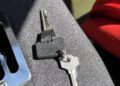 Two metal keys, one with a black plastic head and the other all metal, are lying on a gray fabric surface in bright sunlight—possibly from a 1990 Ferrari F40 offered at auction.