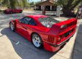 A red 1990 Ferrari F40 sports car is parked on a concrete driveway near vintage gas pumps, with trees and a barn in the background—an iconic scene ready for auction.