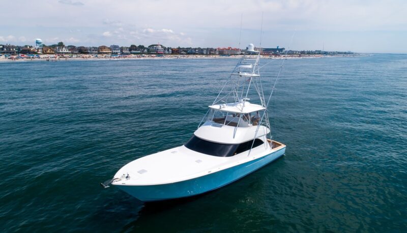 A white and blue sport fishing yacht, exemplifying the ultimate maritime experience, is anchored in calm water near a coastline with a beach and buildings visible in the background.