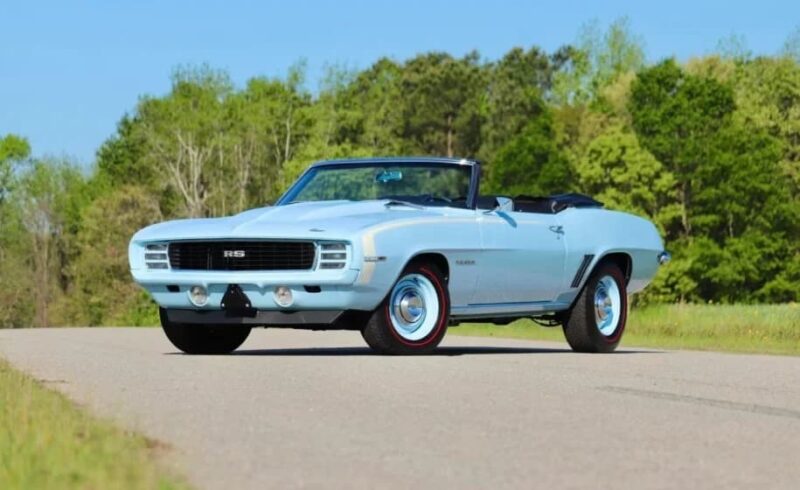 A light blue classic convertible car, likely a late 1960s Chevrolet Camaro RS, is parked on a paved road with green trees in the background—a true icon among high-performance muscle cars.