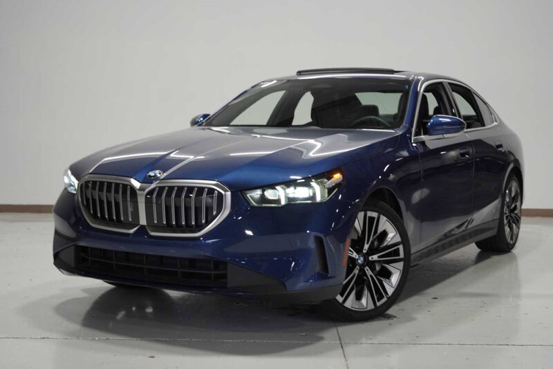 A blue BMW sedan, perfect for the best car lease deals, is parked indoors on a white floor, facing slightly to the left, with headlights on and a visible sunroof.