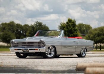 Silver classic convertible car with red interior parked in an empty outdoor lot on a sunny day, perfect for classic cars enthusiasts. Trees and clouds form the scenic background.