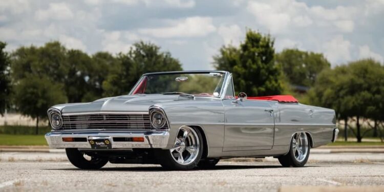 Silver classic convertible car with red interior parked in an empty outdoor lot on a sunny day, perfect for classic cars enthusiasts. Trees and clouds form the scenic background.
