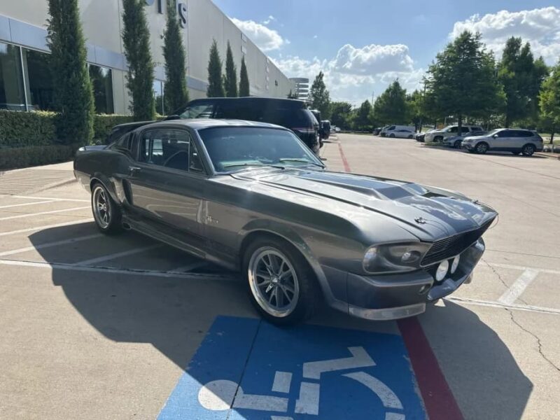 A classic gray Ford Mustang, a standout among classic muscle cars, is parked across two spaces—including a handicap spot—in a parking lot outside a commercial building on a sunny day.