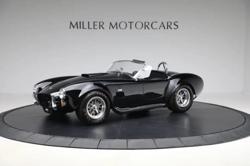 A classic black convertible sports car, a true gem among high-performance cars, is displayed on a circular platform in a showroom with "MILLER MOTORCARS" on the wall behind it.