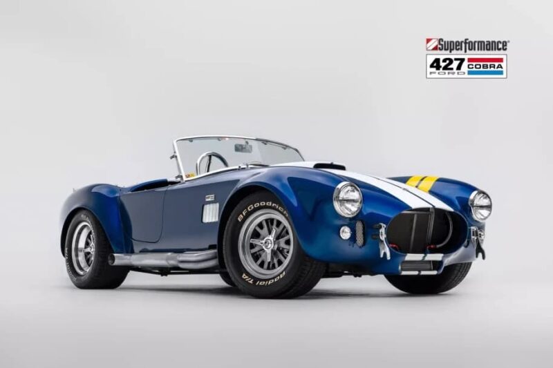 A blue Superformance 427 Ford Cobra sports car with white and yellow racing stripes, shown in a studio setting with a white background—an iconic choice for Classic Cars enthusiasts.