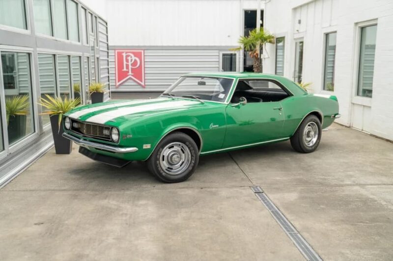 A green classic Chevrolet Camaro Z/28 coupe, a true icon among Classic Muscle Cars, is parked on a concrete driveway near modern glass and white-paneled buildings.