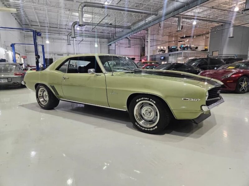 A green 1969 Chevrolet Camaro coupe, a true icon among classic muscle cars, is parked in a brightly lit indoor showroom with several other cars in the background.