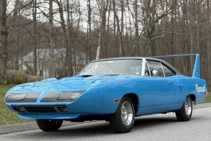 A blue vintage Plymouth Superbird, a true icon among classic muscle cars, is parked on a road with trees and a house in the background.