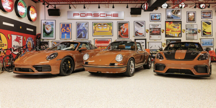 Three Sepia Brown Porsches, celebrating 50 Years of automotive excellence, are parked in a garage filled with collector memorabilia, car-themed posters, and vibrant neon signs.