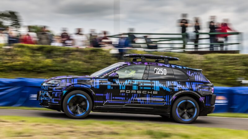 An All-Electric Cayenne with a camouflage wrap drives past spectators on the Shelsley Walsh Hill Climb, chasing an SUV record as motion blur streaks through the background.