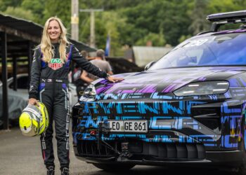 A woman in a racing suit stands next to an All-Electric Cayenne sports car, holding a helmet, with greenery and other vehicles in the background at the Shelsley Walsh Hill Climb.
