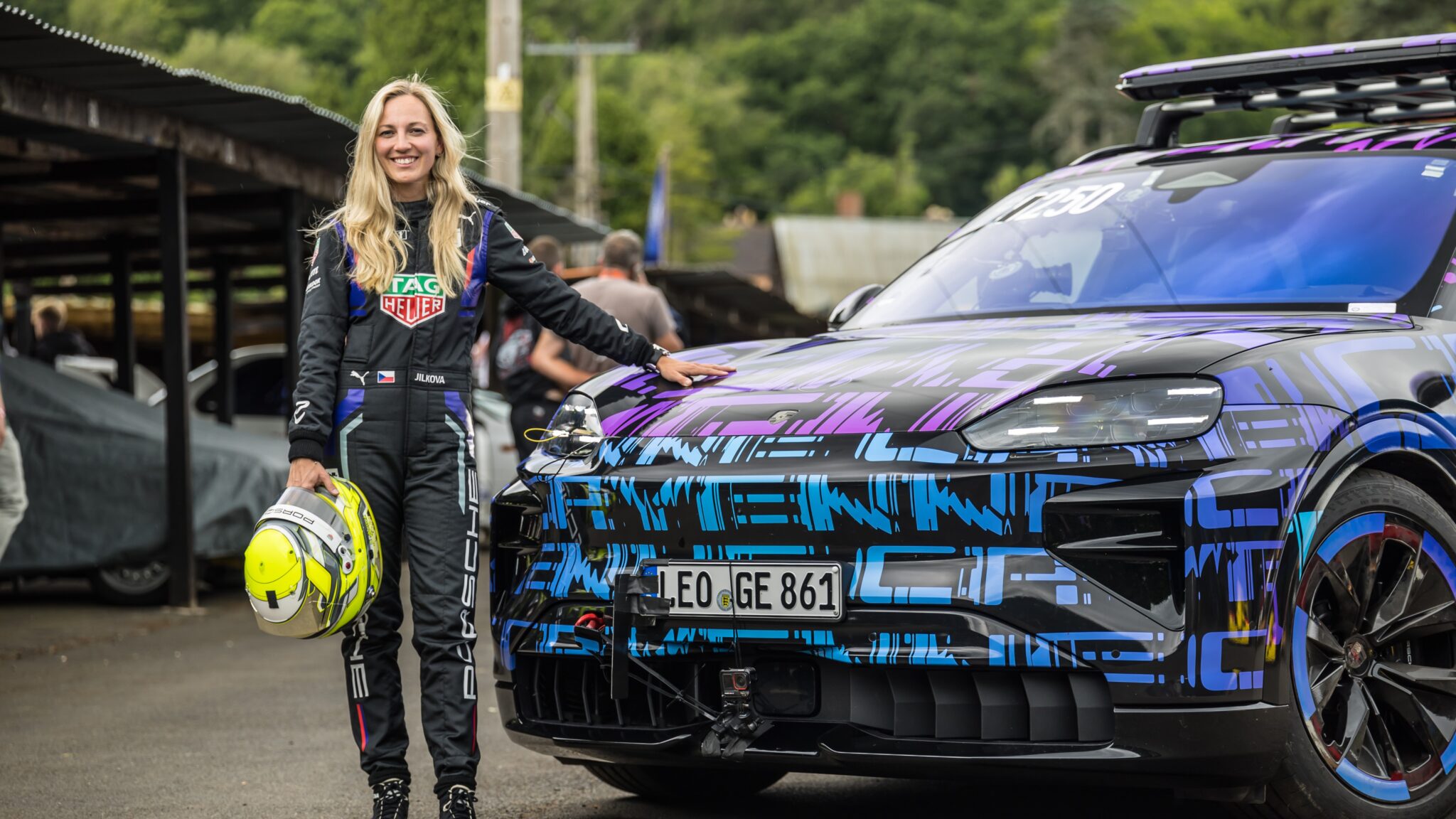 A woman in a racing suit stands next to an All-Electric Cayenne sports car, holding a helmet, with greenery and other vehicles in the background at the Shelsley Walsh Hill Climb.