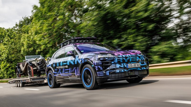 A camouflaged Porsche All-Electric Cayenne SUV tows a classic car on a trailer down a road, surrounded by greenery and blurred trees in the background—hinting at possible SUV record runs like those at Shelsley Walsh Hill Climb.
