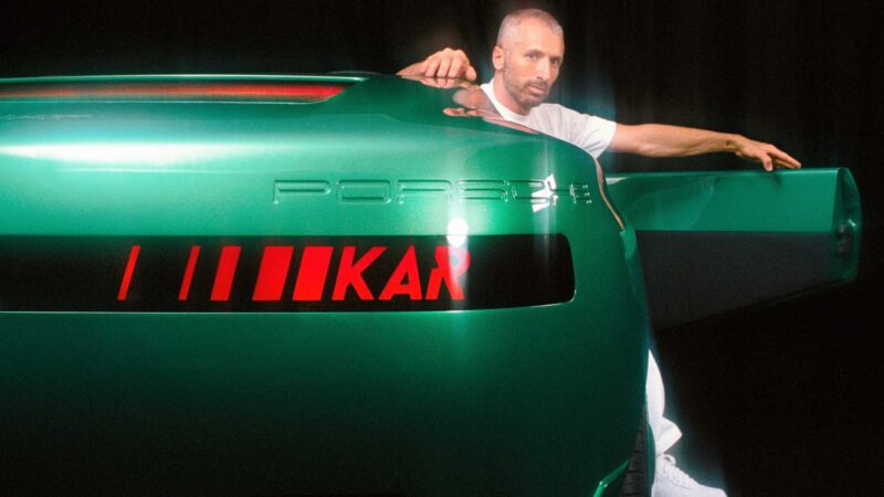 A man in a white t-shirt leans on the open door of a green Porsche Carrera GT with “KAR” and red stripes on the rear, evoking the spirit of a tribute capsule collection.