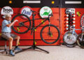 A collector in shorts and a cap repairs a bicycle on a stand in a garage decorated with vintage oil and gas signs, surrounded by sepia brown Porsches from the past 50 years.