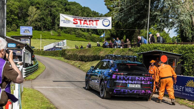 An All-Electric Cayenne waits at the start line of the Shelsley Walsh Hill Climb, with two marshals in orange suits nearby and spectators watching from the sidelines, hoping to witness a new SUV record.