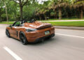 A Sepia Brown Porsche convertible, a collector's favorite for over 50 years, drives down a tree-lined road in daylight.