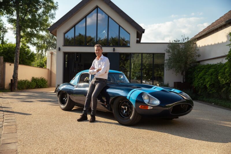 A man stands next to a vintage black Jaguar E-Type with blue stripes, parked in front of a modern house on a sunny day—a scene worthy of Petrolicious’s Film Friday.