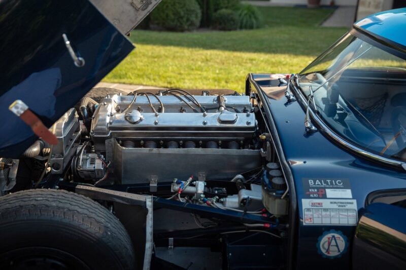 Close-up view of a Jaguar E-Type’s engine bay with the hood open, revealing detailed components and wiring; part of the classic car's exterior and tire are also visible—perfect for any Petrolicious or Film Friday enthusiast.