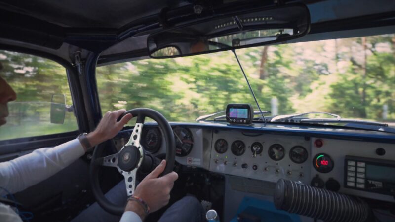 View from the passenger seat of a Jaguar E-Type, showing the driver’s hands on the wheel, dashboard gauges, and a GPS device, with blurred greenery outside—perfect for a classic Petrolicious Film Friday moment.