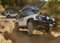 A white off-road vehicle with roof gear navigates a rocky, sandy trail; another similar vehicle is visible in the background among trees and bushes—evoking the spirit of Land Rover Classic to Showcase 10 Historic Expedition Icons at Pebble Beach.