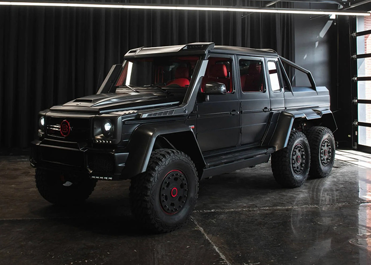 A black six-wheeled Mercedes-AMG G-Wagon with red accents is parked indoors on a polished concrete floor, with black curtains in the background—a true luxury off-road SUV.