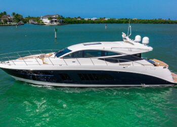 A white and navy blue motor yacht, part of the luxury yachts collection, cruises on calm green water near a shoreline with houses and trees under a clear blue sky.