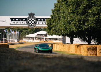 A green sports car drives on a track lined with hay bales at the Goodwood Festival of Speed, with a large event banner and trees in the background.