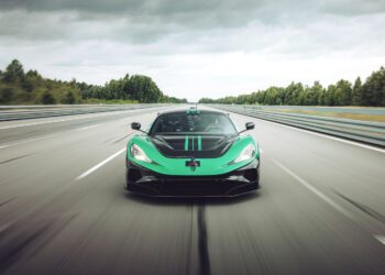 A green and black sports car drives at high speed on an empty highway surrounded by trees under a cloudy sky.