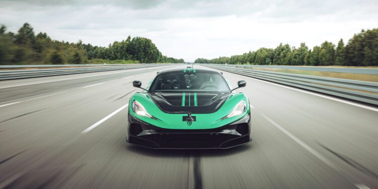 A green and black sports car drives at high speed on an empty highway surrounded by trees under a cloudy sky.
