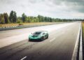 A green Rimac Nevera, the fastest EV known for breaking electric vehicle records, drives alone on a deserted multi-lane highway surrounded by trees under a cloudy sky.
